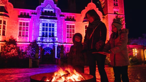 Image shows a family standing around a fire pit with the illuminated hall behind them, at a winter lights event at Blickling Estate, Norfolk